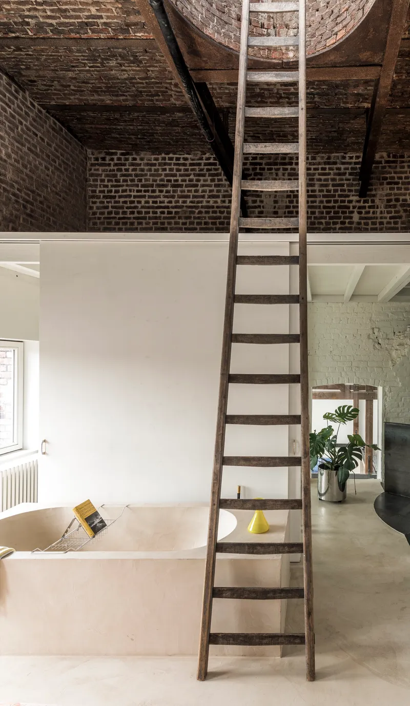 Steel ladder leaning against white partition wall below exposed brick ceiling with timber joists