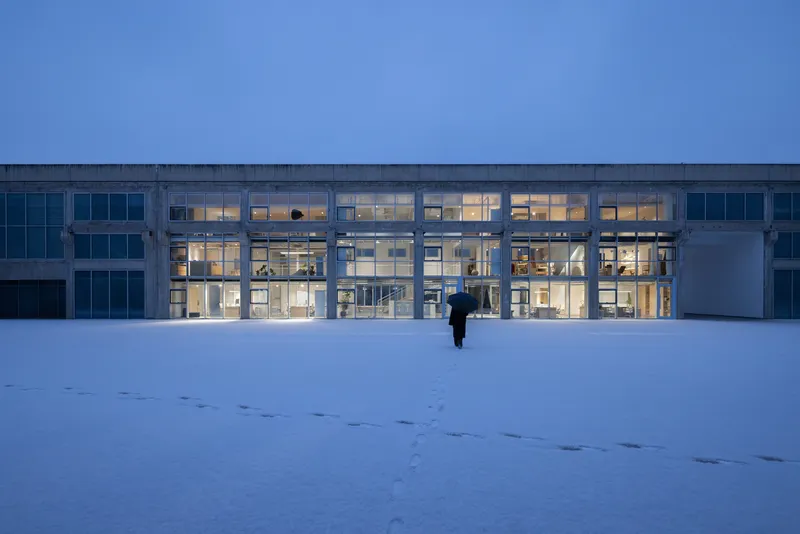 Illuminated glass facade with concrete structural frame viewed across a snow-covered field at dusk