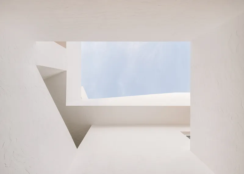 Angular skylight opening carved into white stucco walls framing a view of soft blue sky