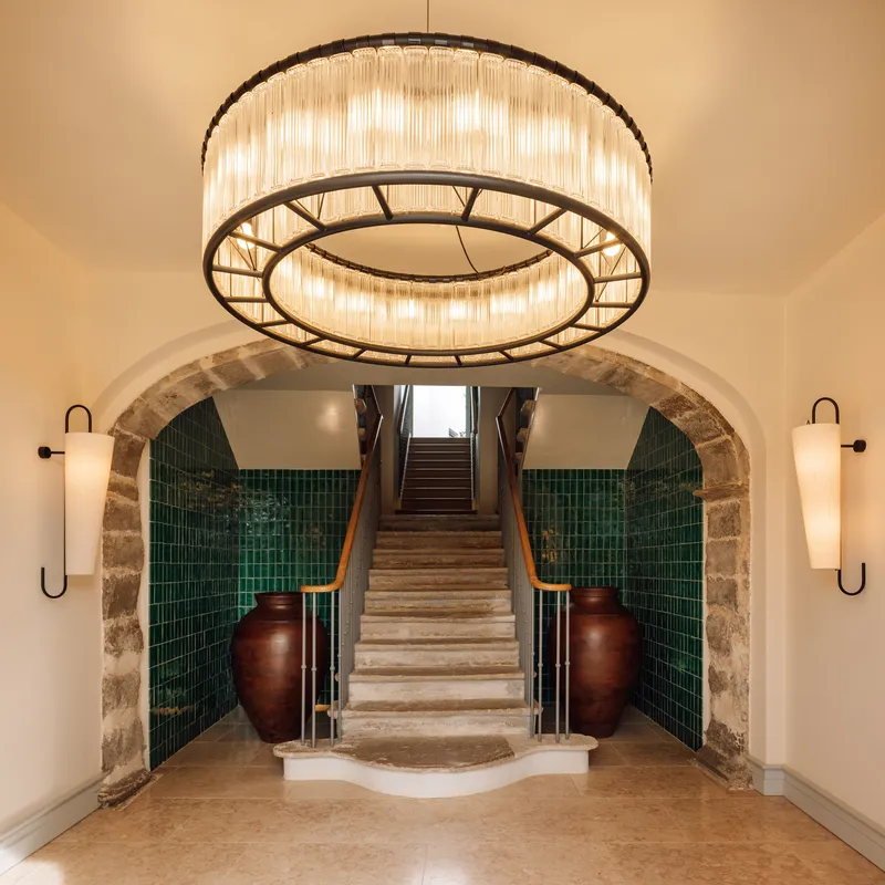 Entry vestibule with stone arch, green tile walls, circular chandelier and marble staircase flanked by ceramic vessels