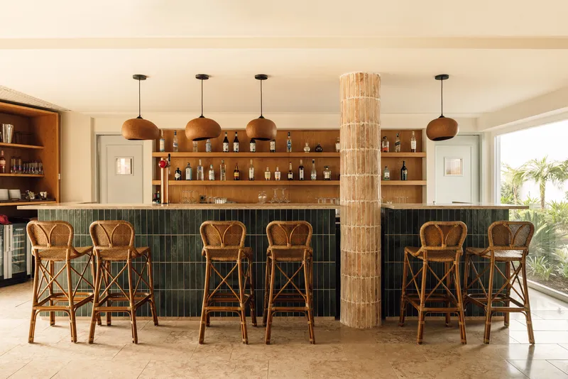 Bar with green tile base, rattan stools, timber shelving and tubular glass column under pendant lights
