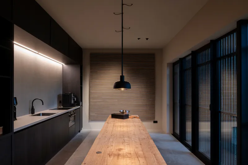 Galley kitchen with dark cabinetry, timber table and horizontal louvered screen illuminated at dusk