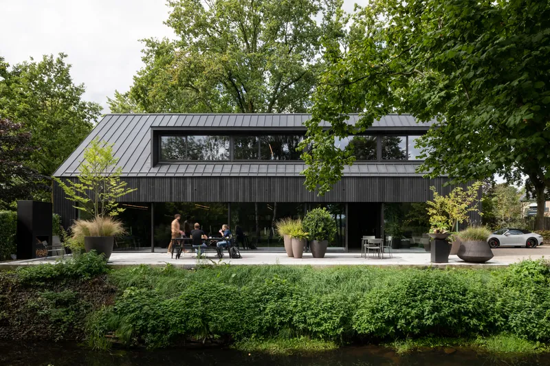 Black timber-clad facade with standing seam metal roof and terrace occupied by people among grasses and planters