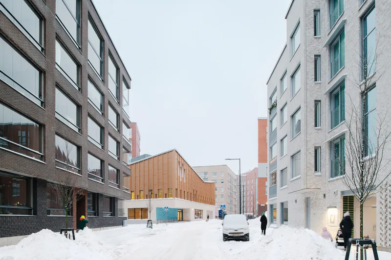 Pedestrian street lined with residential blocks framing the timber-clad volume at the end in heavy snowfall