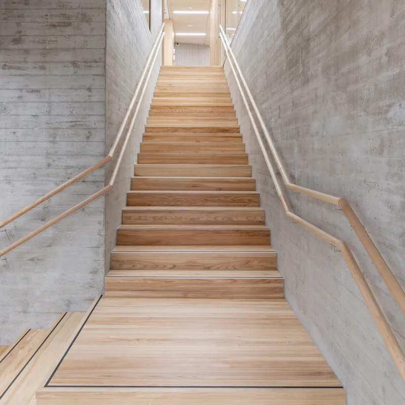 Timber staircase with wood handrails ascending between board-formed concrete walls under natural light