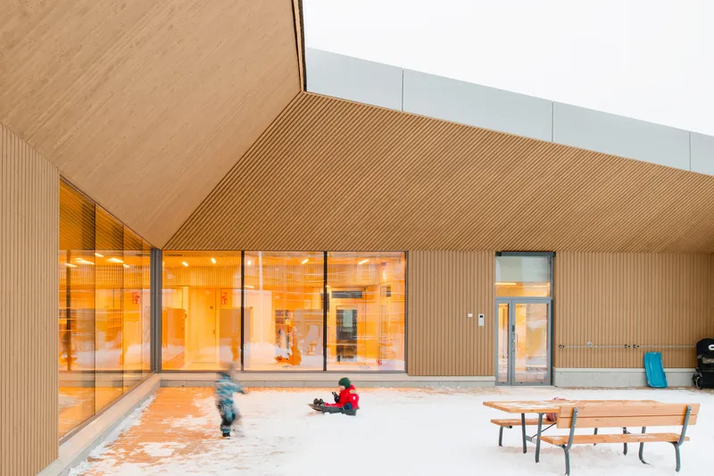 Courtyard with children playing in snow beneath a ribbed wood soffit and backlit orange-glazed windows