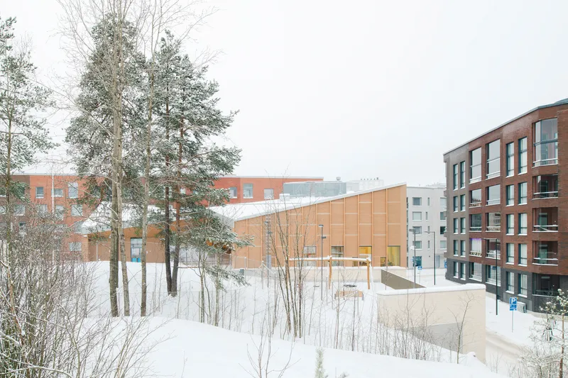 View across snow-covered terrain with pines toward the angled timber-clad volume and surrounding residential blocks