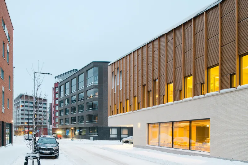 Street view showing the ribbed timber facade above a white brick base in evening snowfall