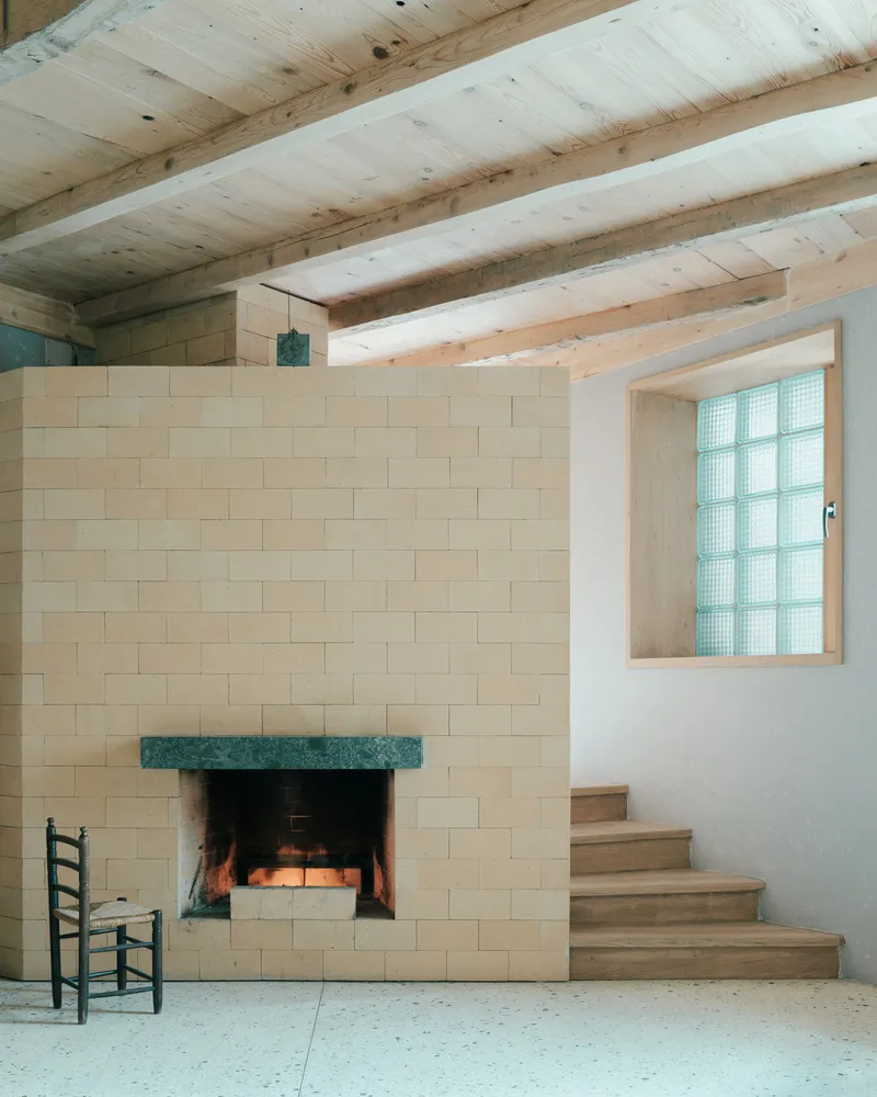 Pale stone fireplace with green marble mantel beneath timber ceiling joists and glass block window