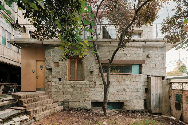 Stone block facade with timber window openings and a young tree in the courtyard below