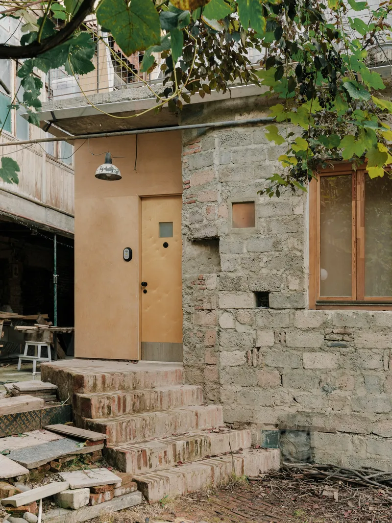 Exterior entry with plywood door and reclaimed brick steps framed by grapevines overhead