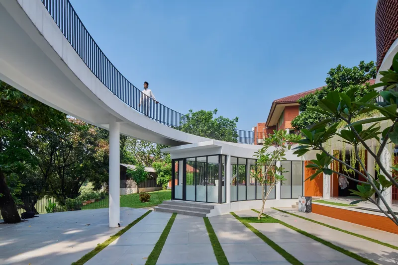 Curving elevated walkway supported by white columns overlooking grass-jointed paving and glazed pavilion