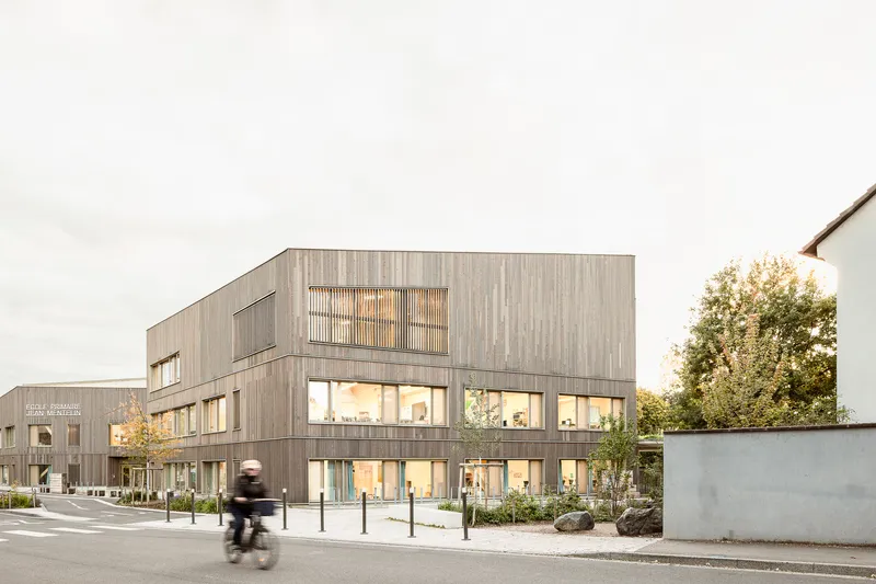 Street view of the timber-clad facade at dusk with a cyclist passing on the road