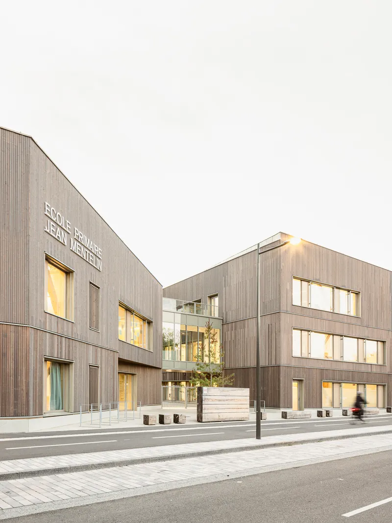 Street corner view at twilight showing two timber volumes with illuminated windows and pedestrians passing