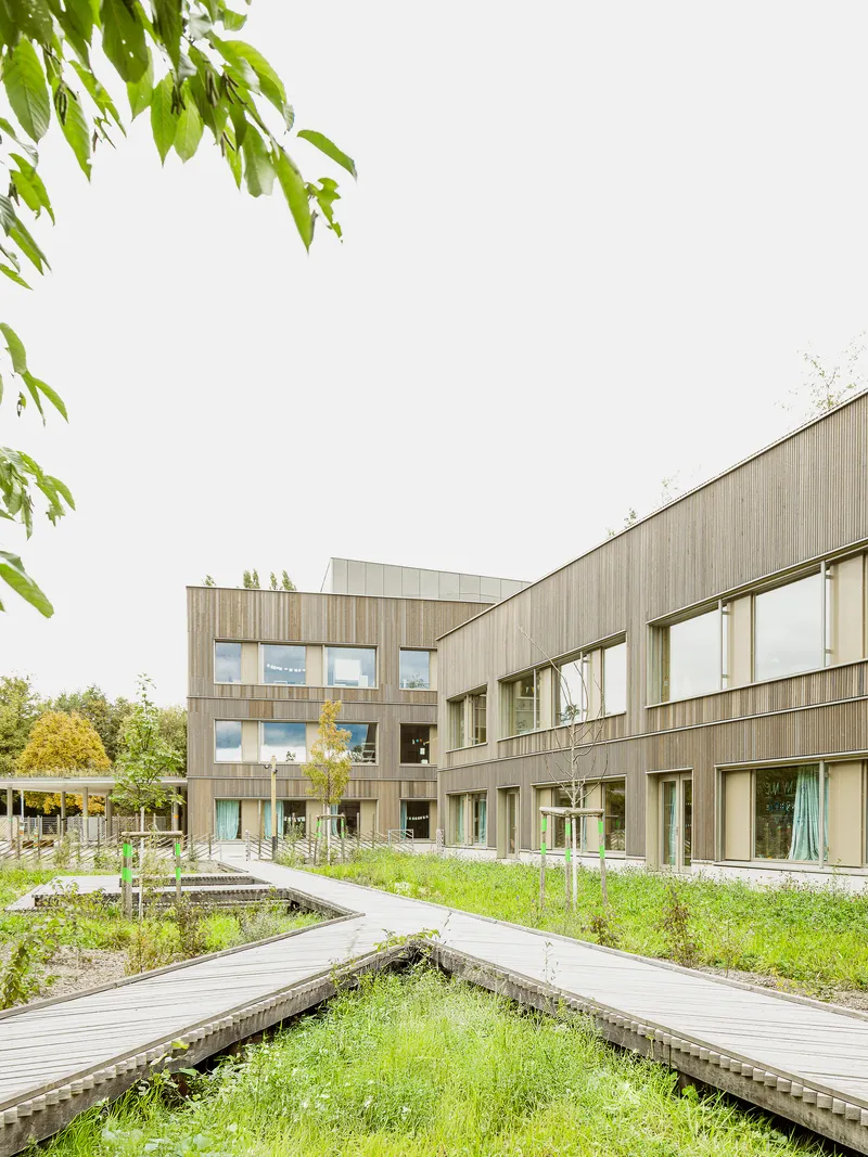 Timber-clad facade with elevated boardwalk paths crossing through planted grasses and wildflowers