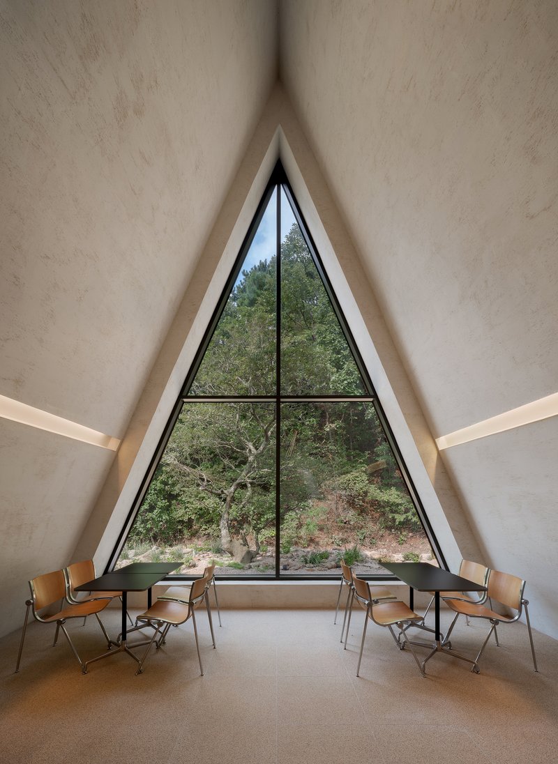 Interior view of a tall triangular window framed in black steel overlooking woodland and chairs