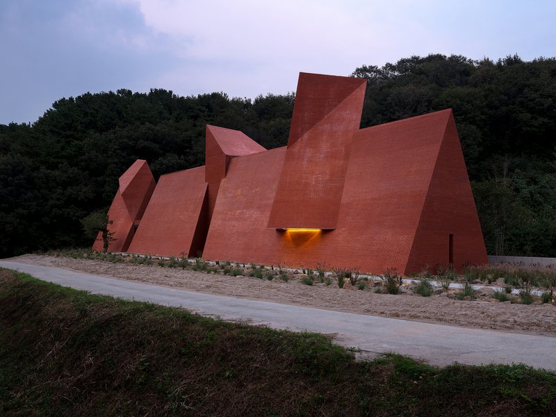 Angular brick facades at dusk with a single illuminated entrance opening beside the roadway