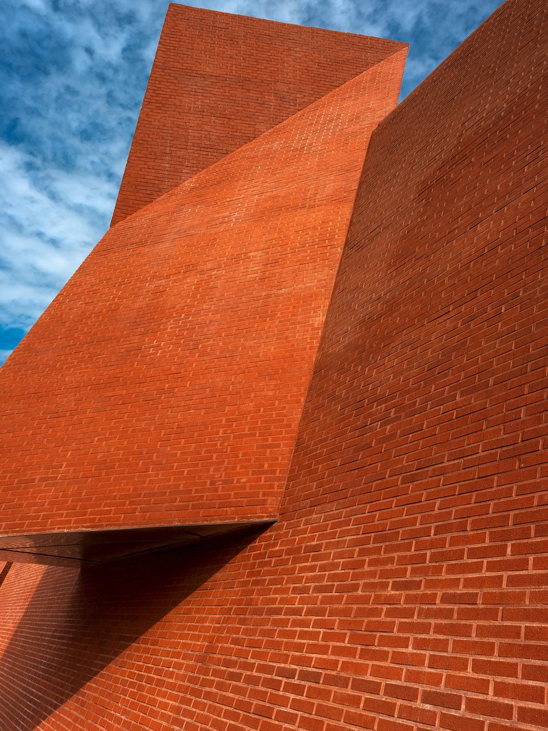 Upward view of folded red brick facade planes meeting at sharp angles under blue sky