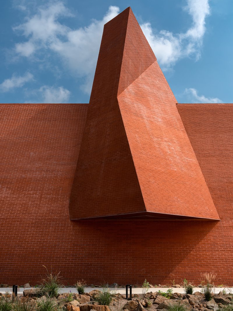 Triangular red brick volume projecting from flat wall above native grasses and rock garden