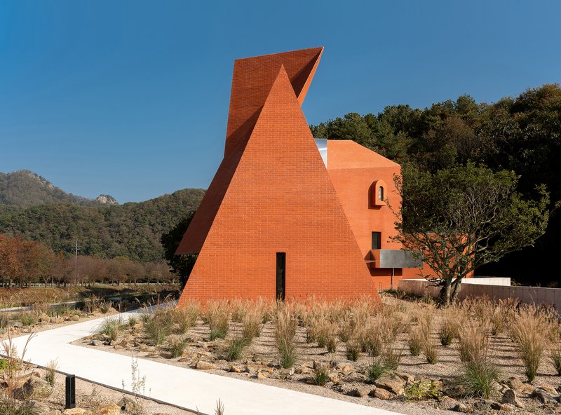 Triangular orange brick volume with a narrow entrance slot set in a planted gravel field
