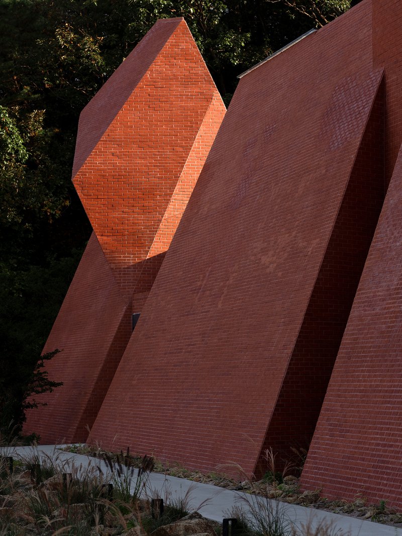 Angled red brick wall with diamond-shaped void catching afternoon sunlight beside native plantings