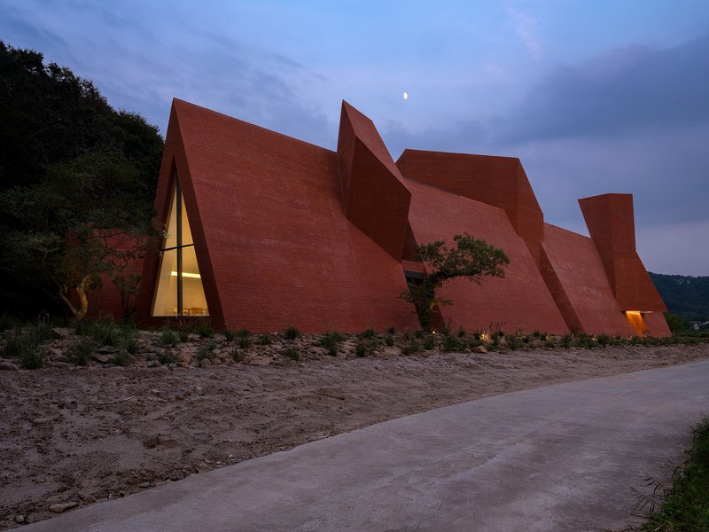 Angular red brick facade with triangular glass openings glowing at twilight beside a concrete path
