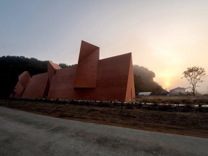 Red geometric volumes with tall triangular forms rising from a gravel landscape at sunrise