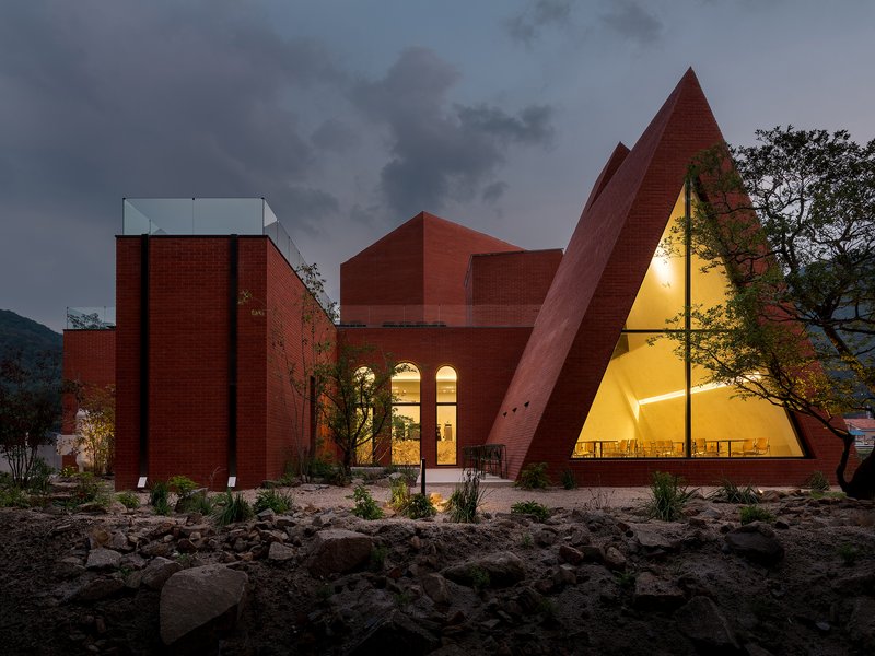 Entrance view showing illuminated triangular glass facade flanked by red brick walls and planted bed