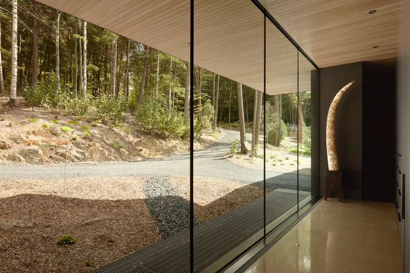 Glass-walled corridor with timber ceiling overlooking a gravel courtyard and forested hillside