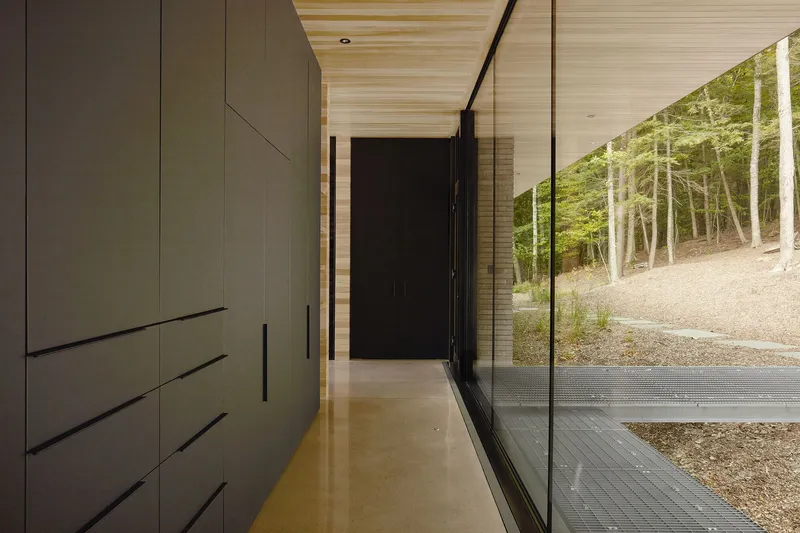 Entry hallway with floor-to-ceiling storage wall opposite frameless glass looking onto gravel and woods