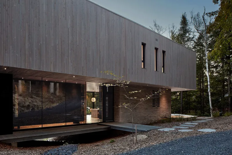 Weathered timber-clad facade with vertical slat windows and covered entry terrace beside a young tree in gravel
