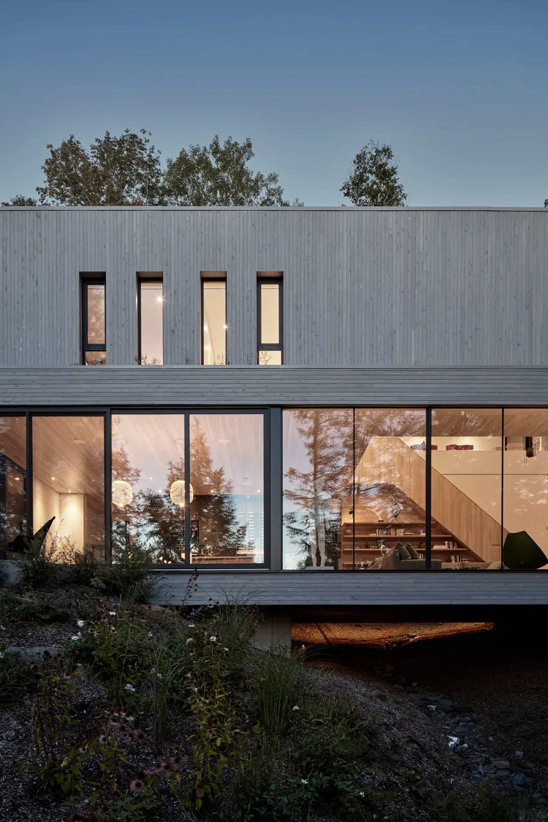 Two-story glazed facade with upper punched windows and lower glass wall reflecting surrounding pines at dusk