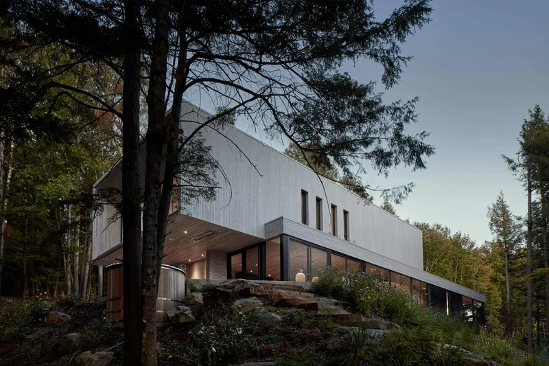 Two-story wood-clad volume on a rocky outcrop framed by tall pine trees at dusk