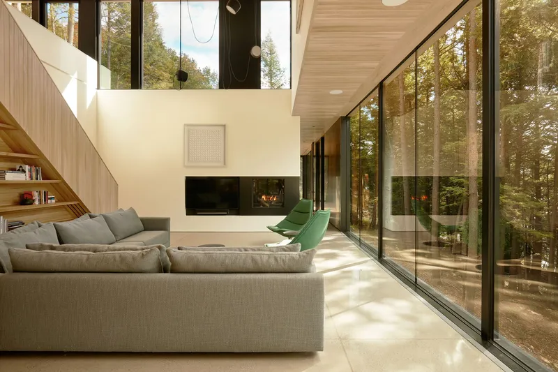 Double-height living room with floor-to-ceiling glazing opening to the forest in afternoon sunlight