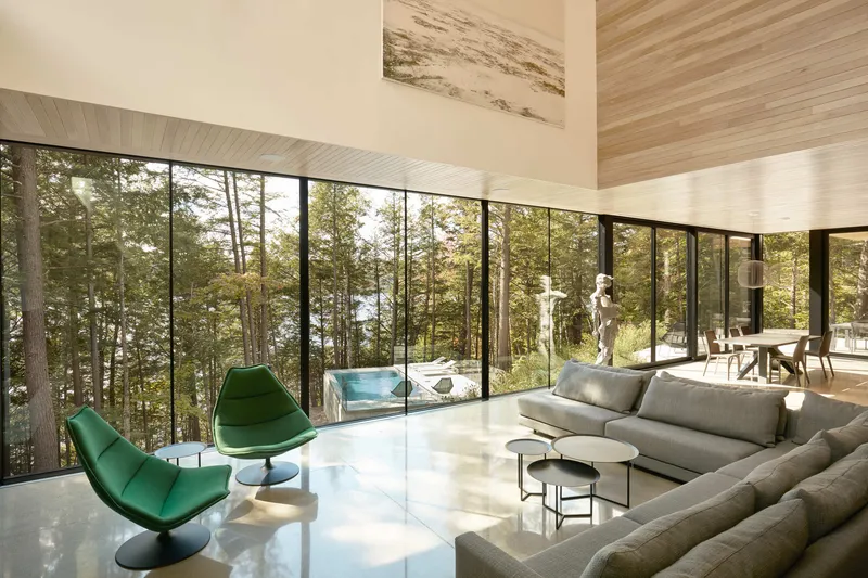 Continuous corner glazing framing views of pine forest with green lounge chairs on polished floor