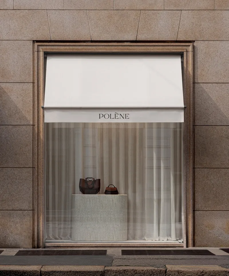 Single window display framed in bronze with white awning and sheer curtains against stone panels