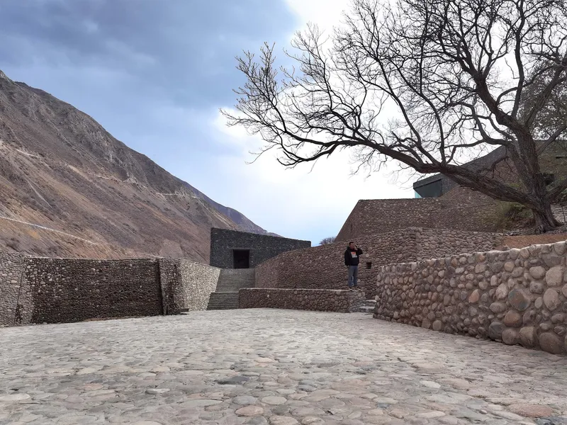 Cobbled courtyard enclosed by stone gabion walls with a figure ascending stairs under a leafless tree