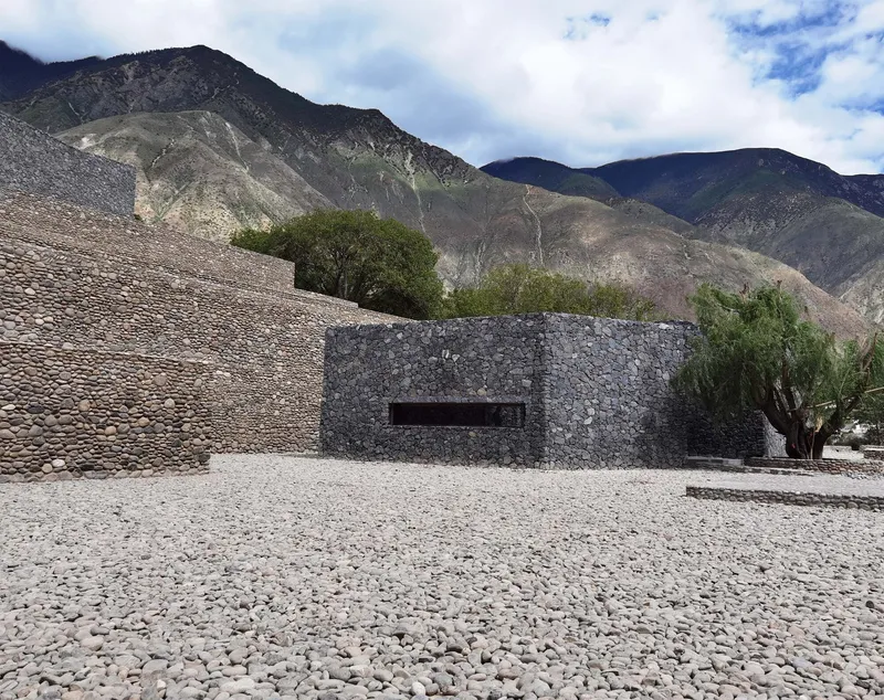 Stone gabion facade with horizontal window opening against a pebbled plaza and mountain backdrop