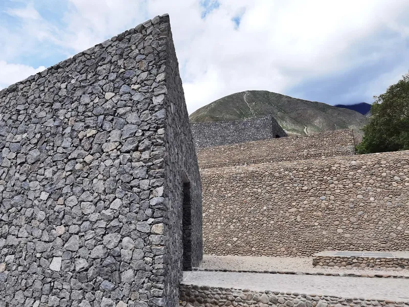 Angled stone wall made of irregular grey rocks set against a hillside under partly cloudy sky