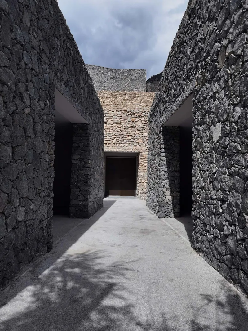 Narrow outdoor passageway between tall stone walls leading to a central opening with dappled shadows
