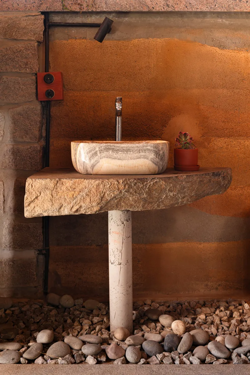 Stone vessel sink on rough-edged counter above river pebble floor with exposed plumbing and terracotta wall