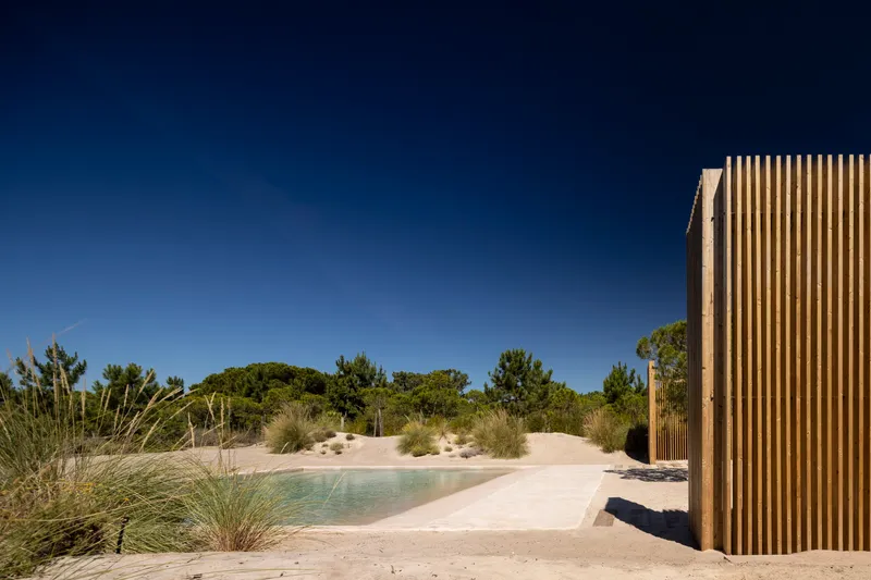 Natural pool edge with sandy beach and dune grasses beneath vertical timber screen under clear sky