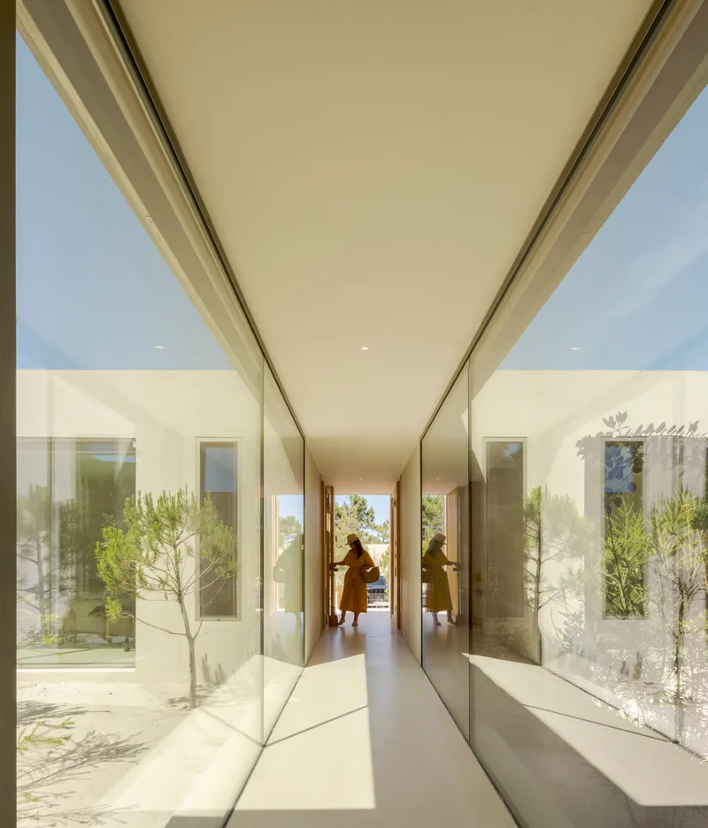 Glass-walled corridor with reflections of courtyard plantings and a figure walking in afternoon light