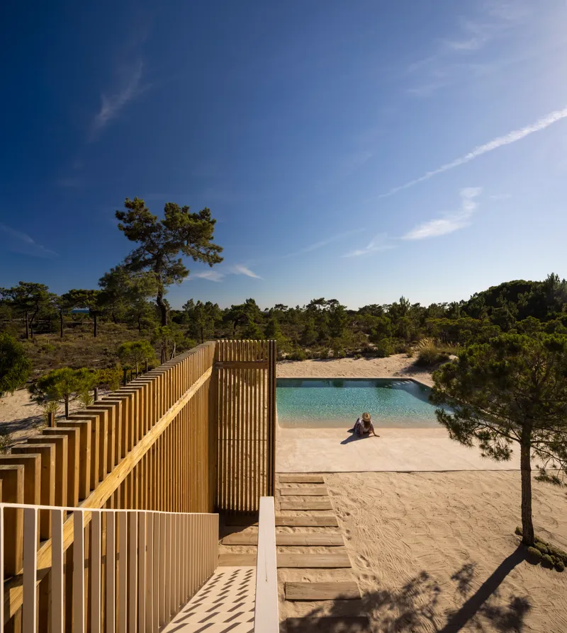 Timber slatted walkway descending toward a pool and sandy beach with pine trees under clear blue sky