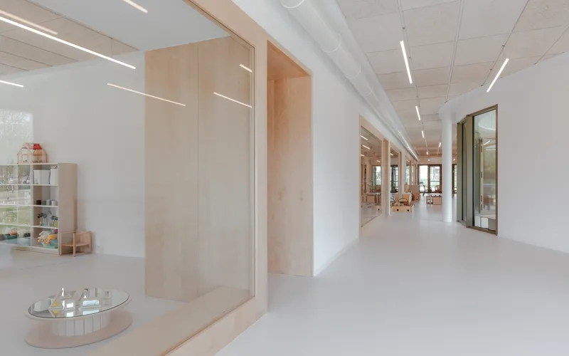 Corridor with pale resin floor, white walls, and plywood-framed openings beneath linear ceiling lights