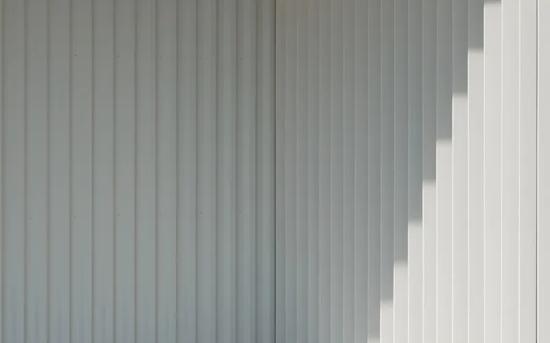 Close-up of vertical corrugated metal cladding in light grey with diagonal sunlight shadows across the surface