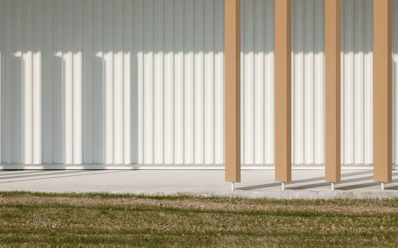 Timber colonnade with square columns casting sharp shadows onto lawn and corrugated metal facade behind