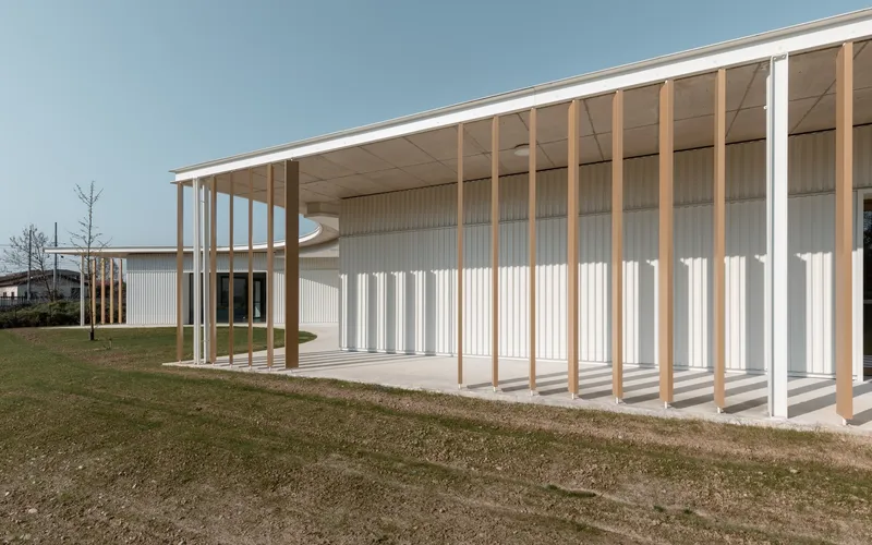 Covered walkway with timber columns and exposed plywood ceiling framing the corrugated facade and lawn in late afternoon