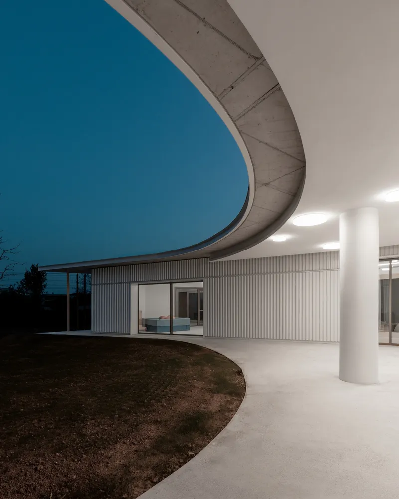 Covered exterior porch with curved concrete canopy, white columns and vertical metal cladding at dusk
