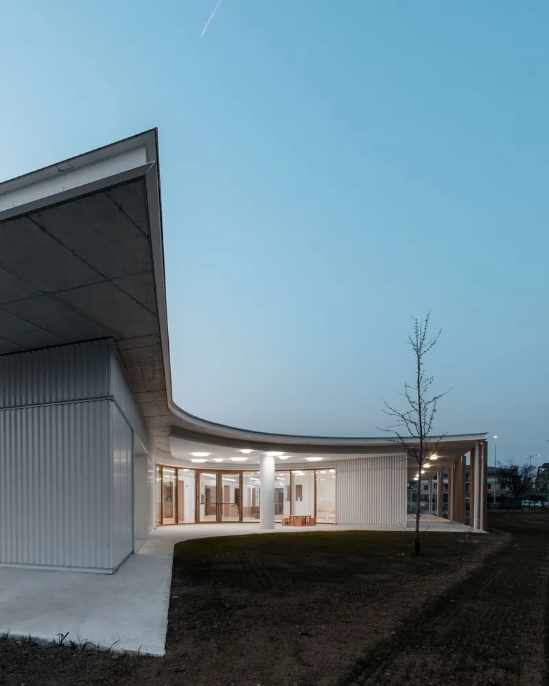 Exterior view showing the curving metal-clad facade and concrete roof overhang with a young tree at twilight